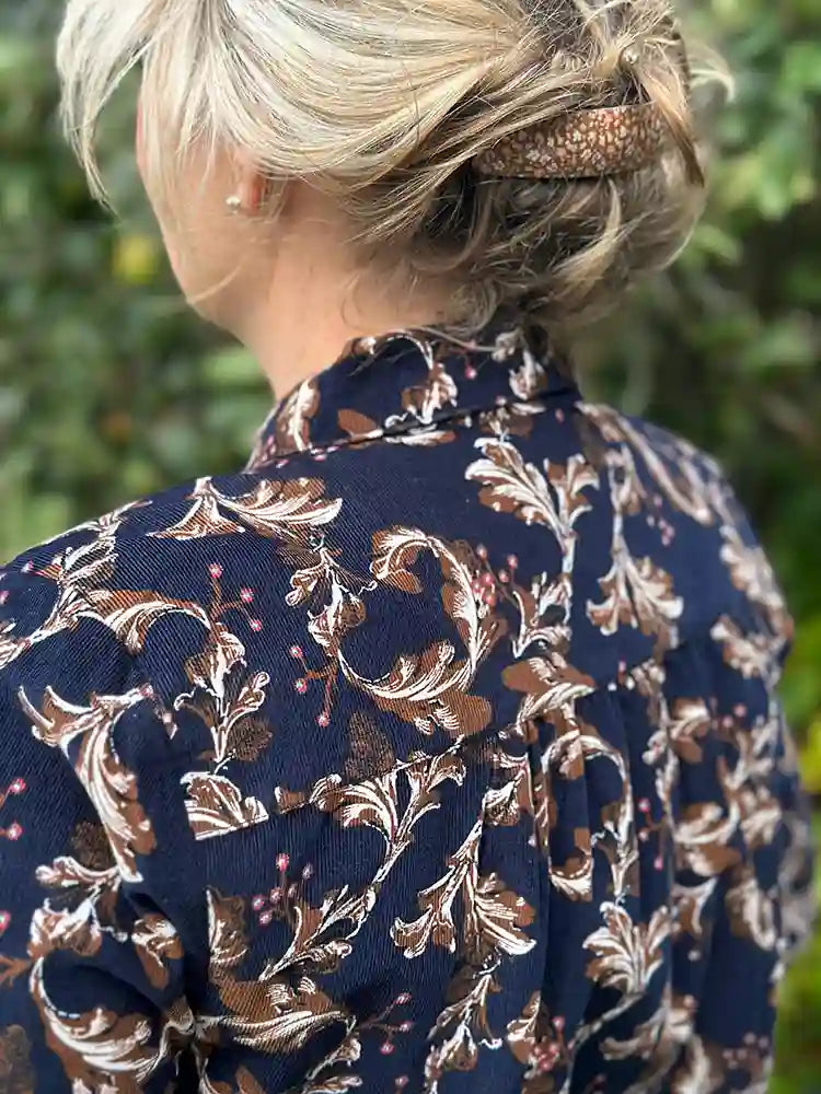 A woman shown from the back wearing a navy blue blouse with a brown and beige botanical pattern, styled with a textured brown hair accessory, set against a soft-focus green outdoor background.
