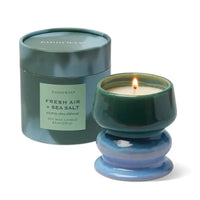 A Paddywax soy wax candle in a blue and green ceramic pedestal holder next to its matching round tin packaging on a white background.