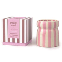 A pink and cream striped ceramic candle holder next to its matching striped packaging box, labeled 'grecian sand' scented soy wax candle.