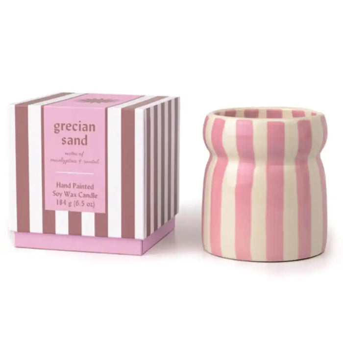 A pink and cream striped ceramic candle holder next to its matching striped packaging box, labeled 'grecian sand' scented soy wax candle.