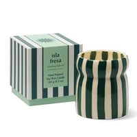 A hand-painted soy wax candle in a ribbed ceramic pot with dark green and cream vertical stripes, shown next to its matching striped packaging box labeled 'isla fresa'.
