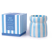 A hand-painted soy wax candle in a ribbed cylindrical pot with light blue and white vertical stripes, shown next to its matching striped packaging box labeled 'lost at sea'.