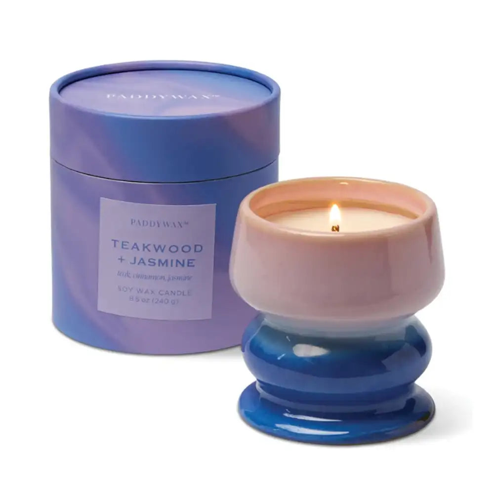 A lit Paddywax soy wax candle in a ceramic holder with a blue-to-pink gradient base, shown next to its matching blue-purple cylindrical packaging container on a white background.