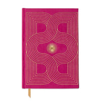 A bright pink hardcover notebook with a gold geometric pattern and a small gold sunburst emblem on the cover, featuring a gold ribbon bookmark.