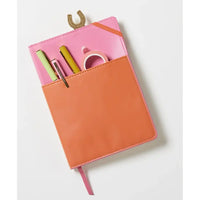 A pink and orange zip-around notebook or planner cover shown open on a light gray background, with pens, tape, and a gold horseshoe-shaped accessory tucked into the pockets.