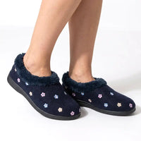 A person wearing dark navy slip-on slippers with a fuzzy navy trim and small embroidered multicolor flowers, shown against a light gray studio background.
