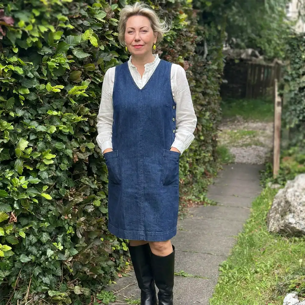 Blonde model facing the camera with lime green earrings wearing a white long sleeve blouse and denim pinafore dress. Hand in pinafore dress pockets and black knee high boots on. Stood on garden path with greenery surrounding her. 