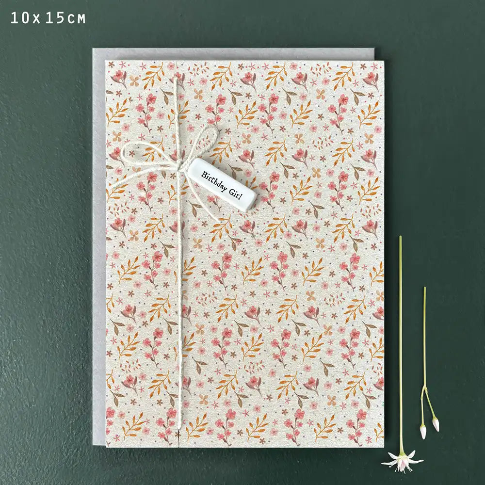 Pale ecru coloured card with delicate floral pattern in mustards, pinks and greens. White string down the left hand side tied into a bow with a white porcelain tag. Porcelain tag with black delicate writing with the words 'Birthday Girl'. 