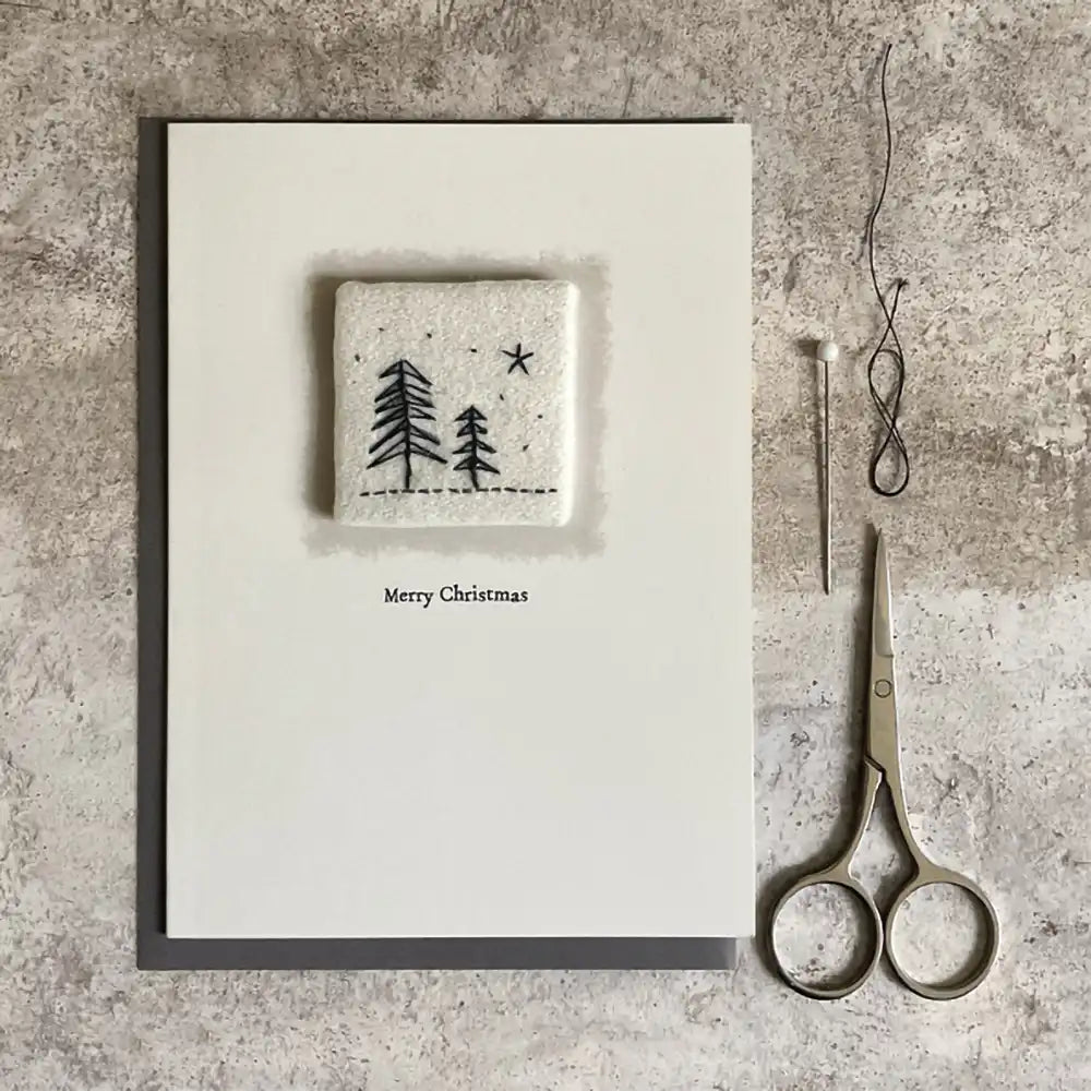 East of India embroidered Christmas card. Embroidered patch on a plain cream card with a simply 'Merry Christmas' greeting. Embroidery of trees and stars. Shown on a stone surface with a pin and scissors for scale.