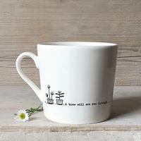 White mug with line drawings of plants and the message 'A brew will see you through...' Pictured against a rough light wooden background and a daisy lying by the cup.