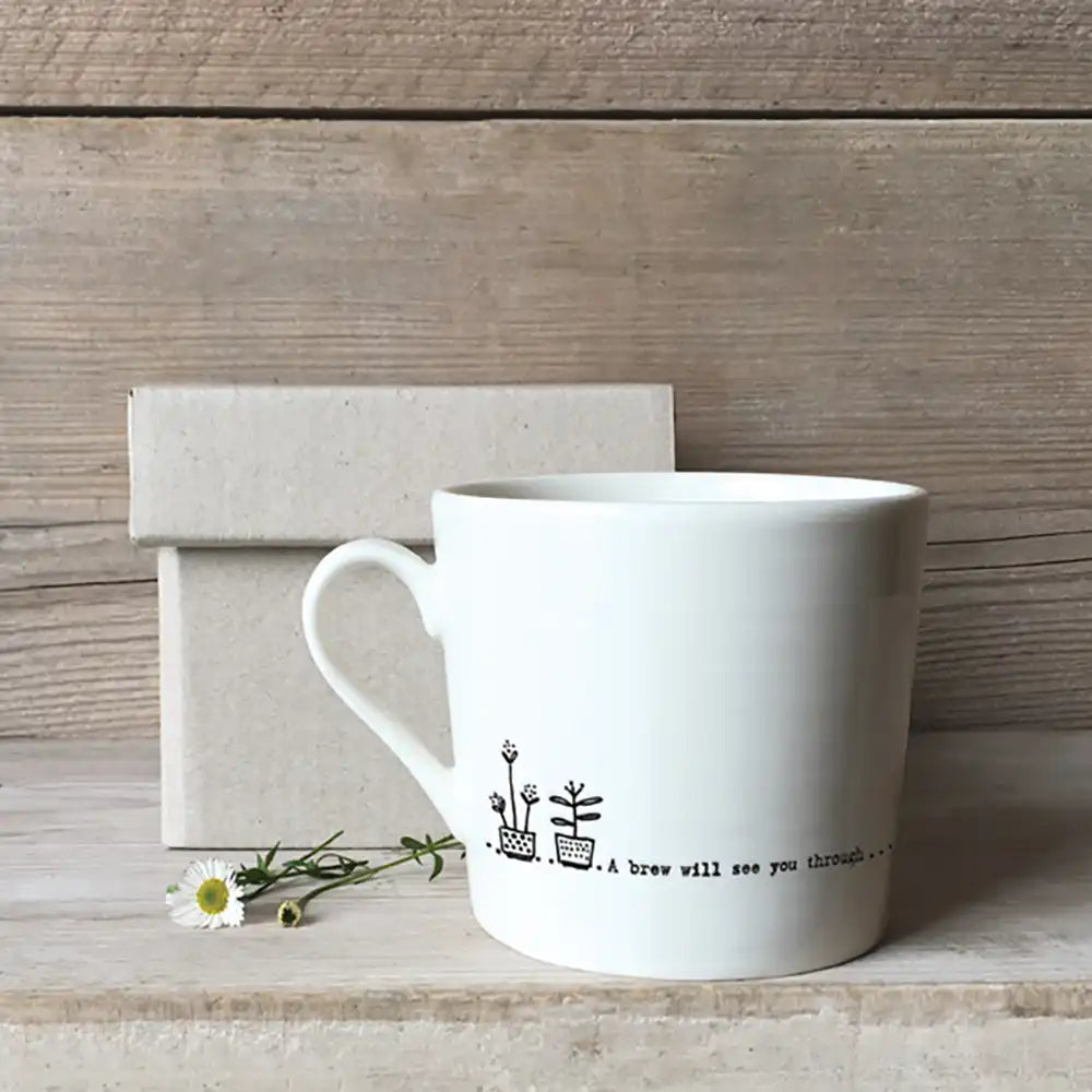 White mug with line drawings of plants and the message 'A brew will see you through...' Pictured against a rough light wooden background, cardboard box and a daisy lying by the cup.