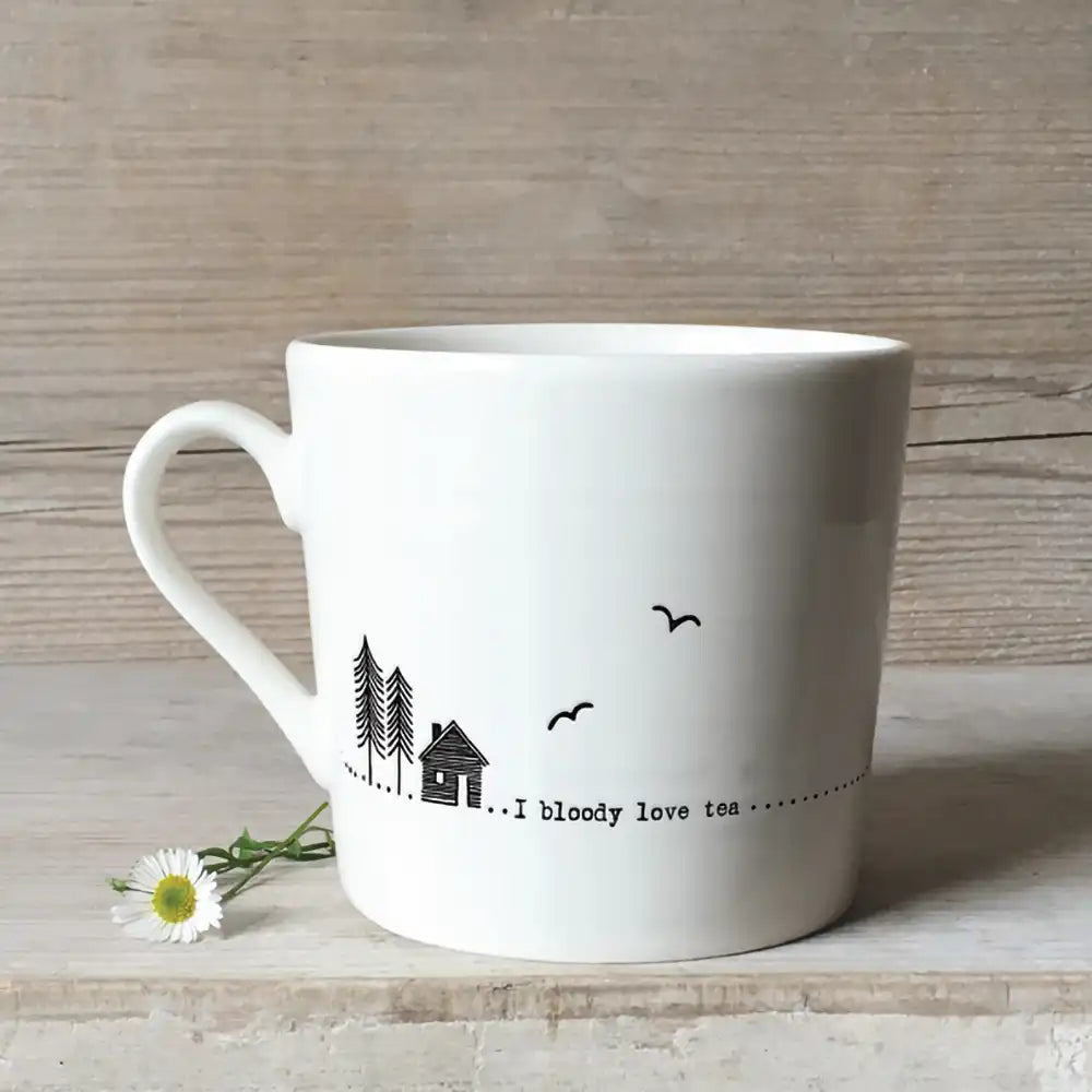 White mug with line drawings of a house and some trees and the message 'I bloody love tea' Pictured against a rough light wooden background and a daisy lying by the cup.