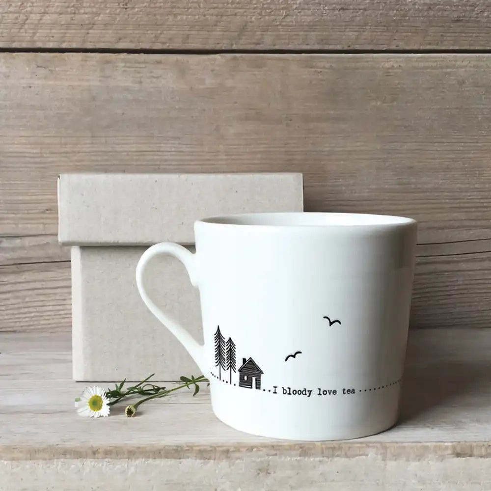 White mug with line drawings of a house and some trees and the message 'I bloody love tea' Pictured against a rough light wooden background a cardboard box and a daisy lying by the cup.