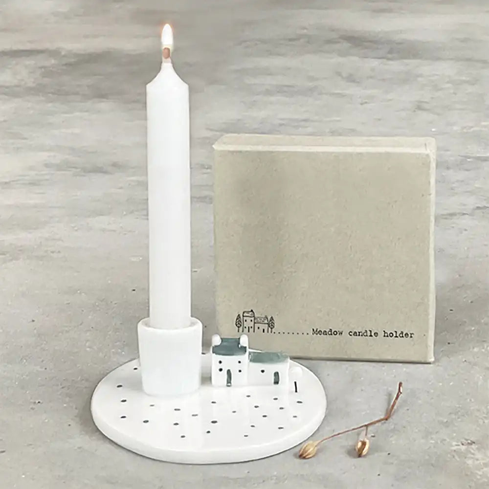 White candle holder with a lit candle on a gray surface, accompanied by a small box labeled 'Meadow candle holder'.