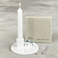 White candle holder with a lit candle on a gray surface, accompanied by a small box labeled 'Meadow candle holder'.