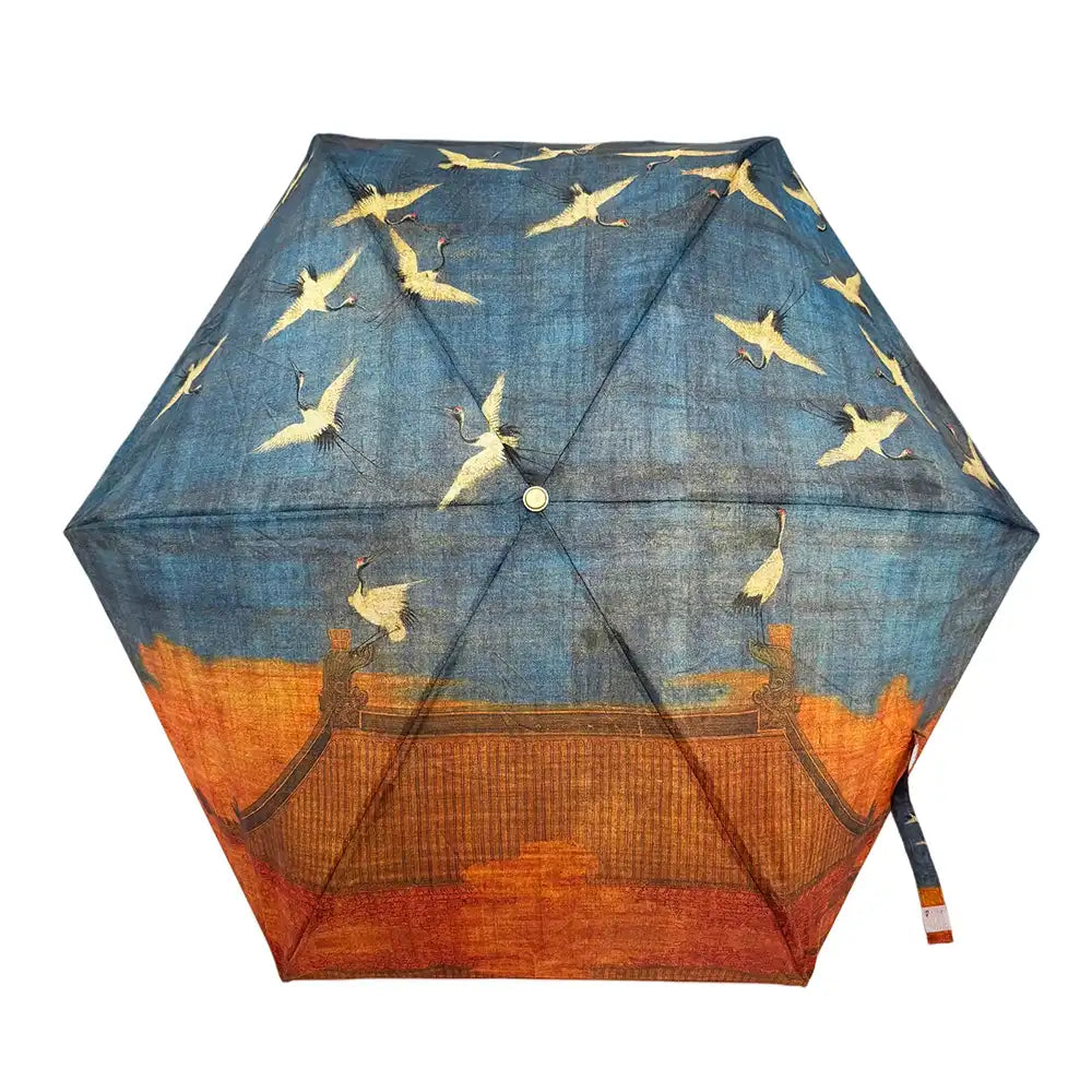 A hexagonal umbrella shown open against a white background, featuring a printed design of white cranes flying across a blue sky above an orange architectural landscape.