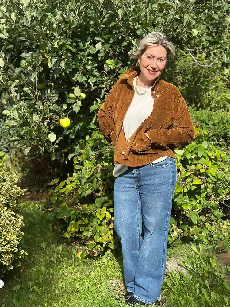 A woman standing outdoors in a garden wearing a brown corduroy jacket over a white top and blue jeans, with leafy green bushes behind her.