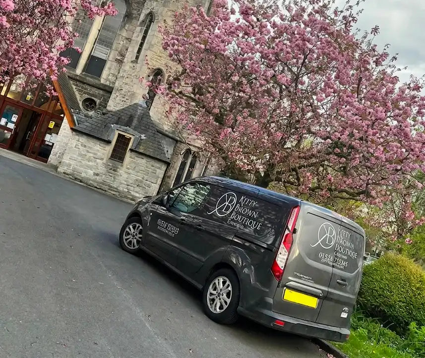 A dark gray Ford Transit van parked on a street in front of a stone building, covered with pink cherry blossoms. The side and rear windows feature white printed branding for 'Bespoke Boutique'.