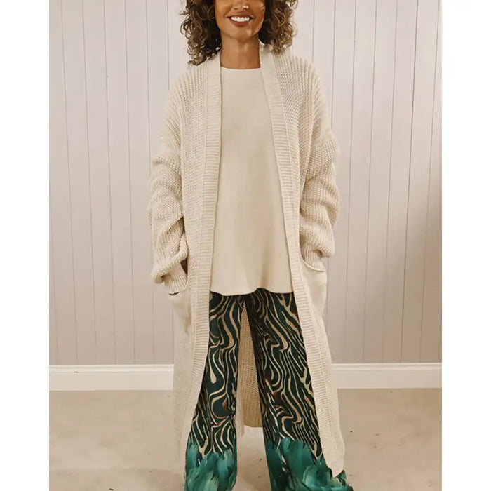 Woman wearing a long beige cardigan, white top, and patterned pants against a neutral background