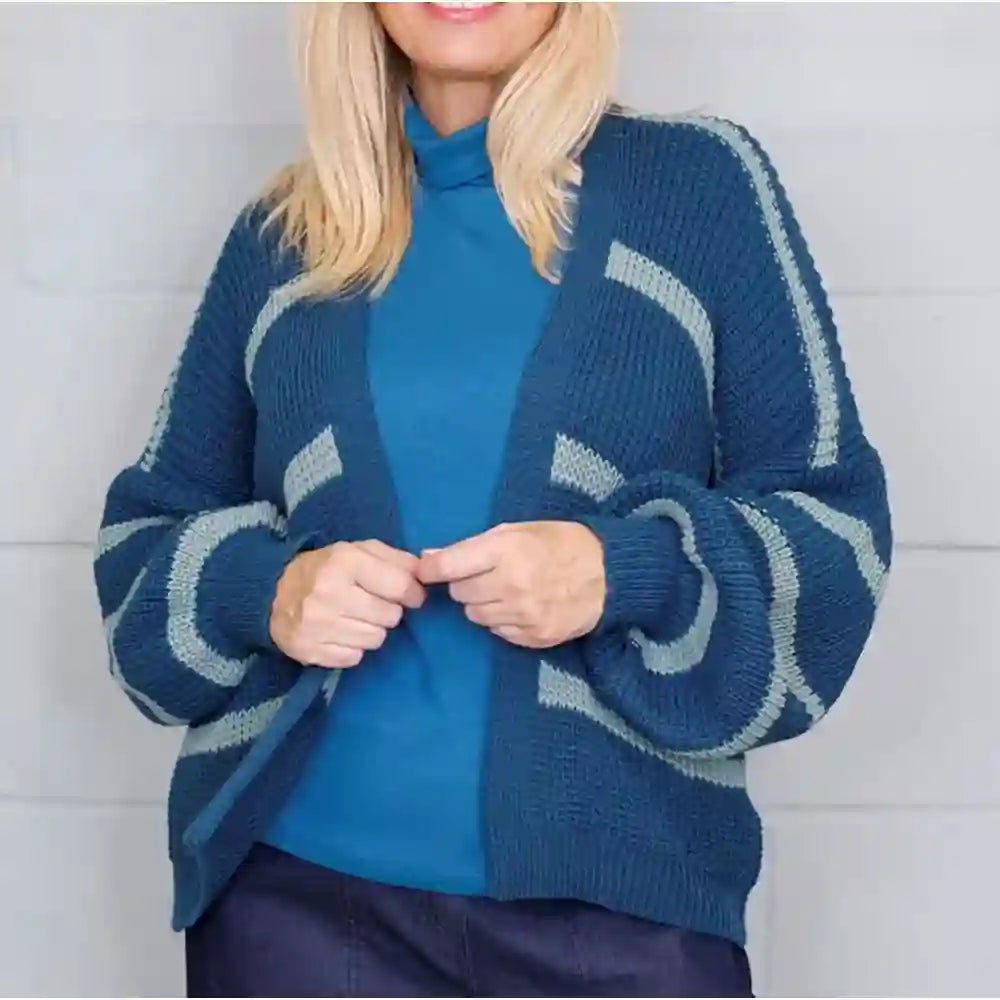 A woman wearing a blue and light gray striped knit cardigan over a solid blue mock-neck top, standing against a light gray paneled wall.