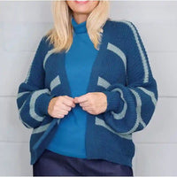 A woman wearing a blue and light gray striped knit cardigan over a solid blue mock-neck top, standing against a light gray paneled wall.