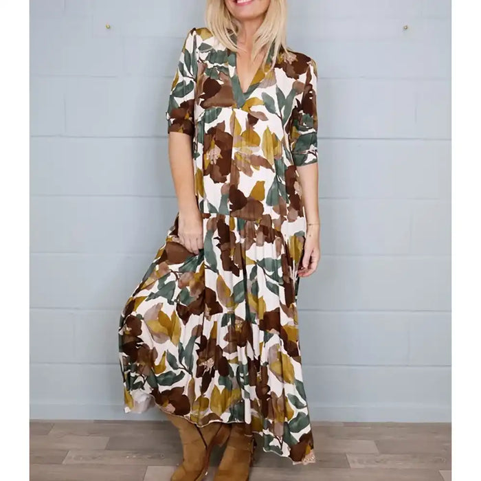 A woman wearing a bohemian-style maxi dress with a brown, olive green, and mustard yellow floral pattern on a cream background, paired with brown suede boots, standing against a light gray paneled wall.