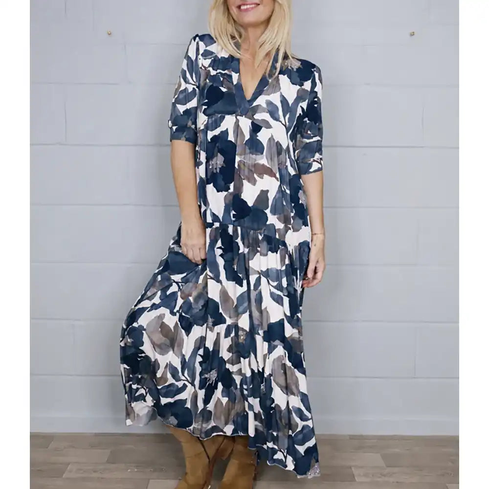 A woman wearing a long-sleeve maxi dress with a navy, gray, and white watercolor floral pattern, standing against a light gray brick wall.