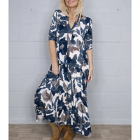 A woman wearing a long-sleeve maxi dress with a navy, gray, and white watercolor floral pattern, standing against a light gray brick wall.