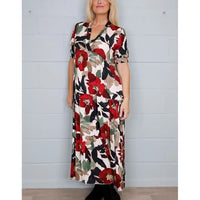 A woman modeling a short-sleeve maxi dress with a bold floral print in red, black, beige, and green on a cream background, standing against a white paneled wall.