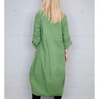 A woman viewed from behind wearing a sage green long-sleeve tunic dress with rolled-up sleeves, standing against a white paneled wall.