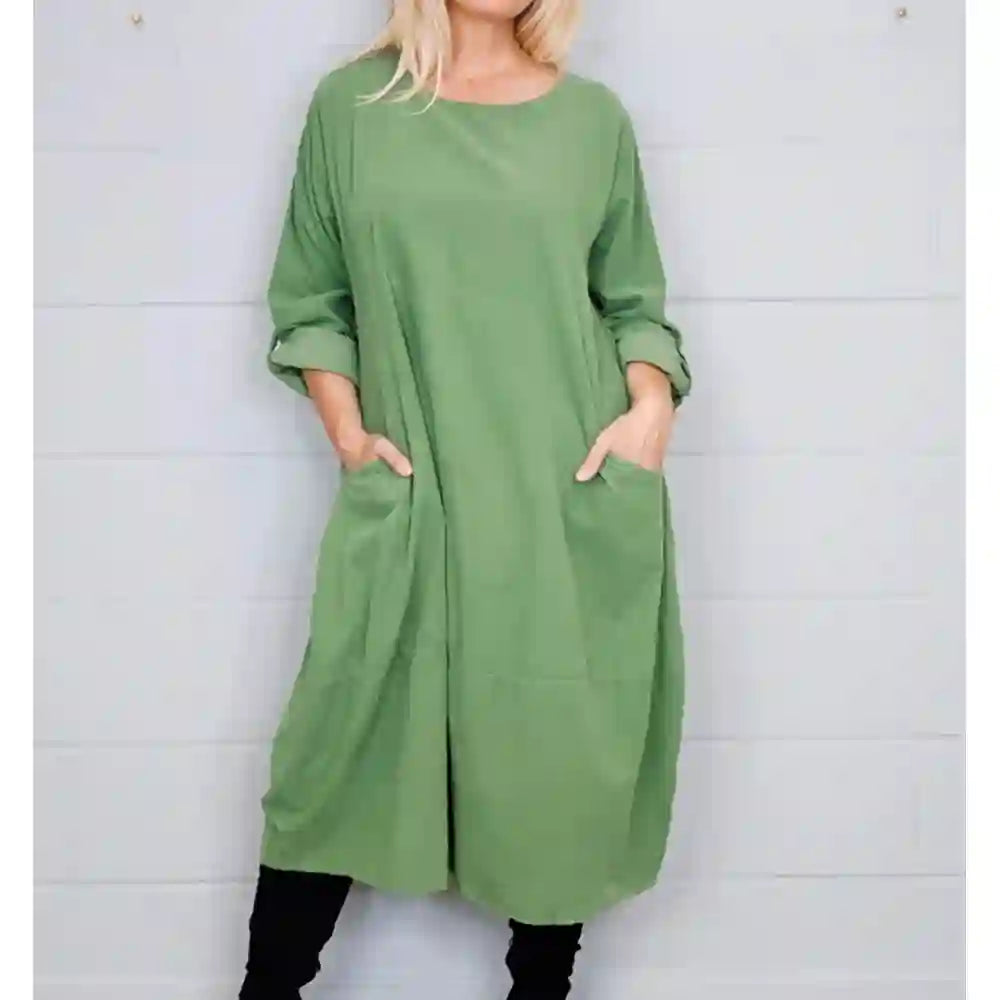 A woman wearing a loose sage green tunic dress with three-quarter sleeves and large front patch pockets, standing against a white paneled wall.