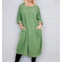 A woman wearing a loose sage green tunic dress with three-quarter sleeves and large front patch pockets, standing against a white paneled wall.