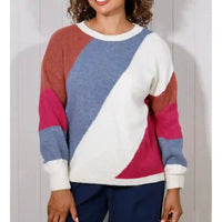 A woman wearing a color-block knit sweater with diagonal panels of white, dusty blue, rust brown, and magenta, paired with navy pants, standing against a white paneled wall.