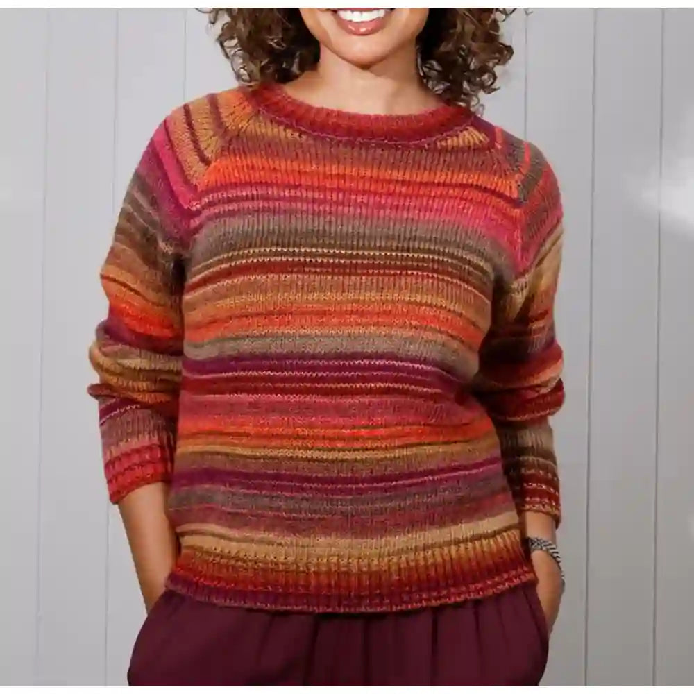 A woman wearing a long-sleeve knit sweater with horizontal stripes in orange, red, pink, brown, and beige, paired with dark purple pants, set against a light gray paneled wall.