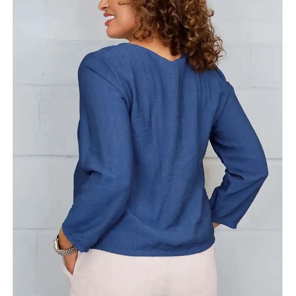 A woman wearing a blue long-sleeve top with vertical pleats and a peplum hem, paired with light beige pants, standing against a white paneled wall.