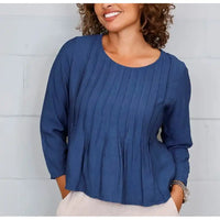 A woman wearing a blue long-sleeve top with vertical pleats and a peplum hem, paired with light beige pants, standing against a white paneled wall.