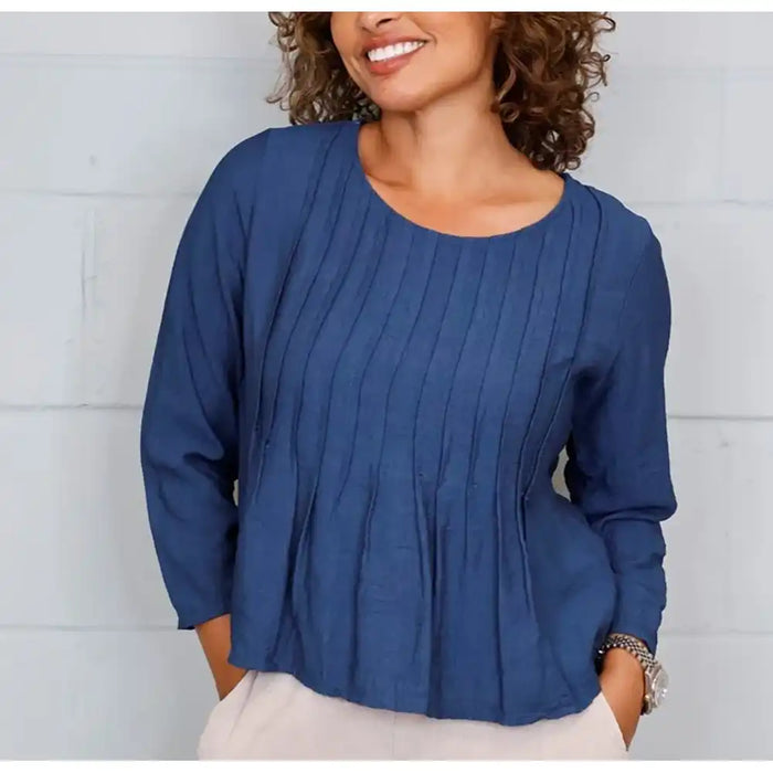 A woman wearing a blue long-sleeve top with vertical pleats and a peplum hem, paired with light beige pants, standing against a white paneled wall.