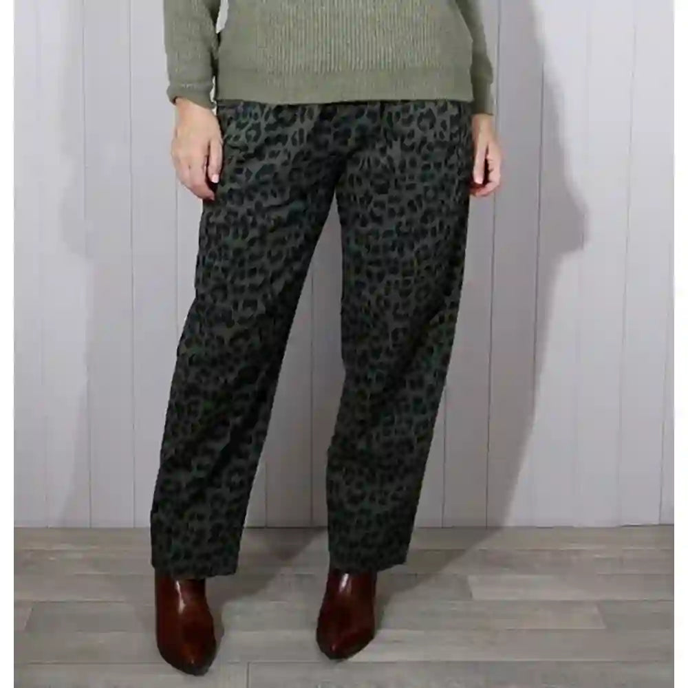 A person wearing dark green and black leopard-print trousers, paired with an olive green sweater and brown pointed-toe boots, standing against a light gray paneled wall.