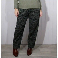 A person wearing dark green and black leopard-print trousers, paired with an olive green sweater and brown pointed-toe boots, standing against a light gray paneled wall.