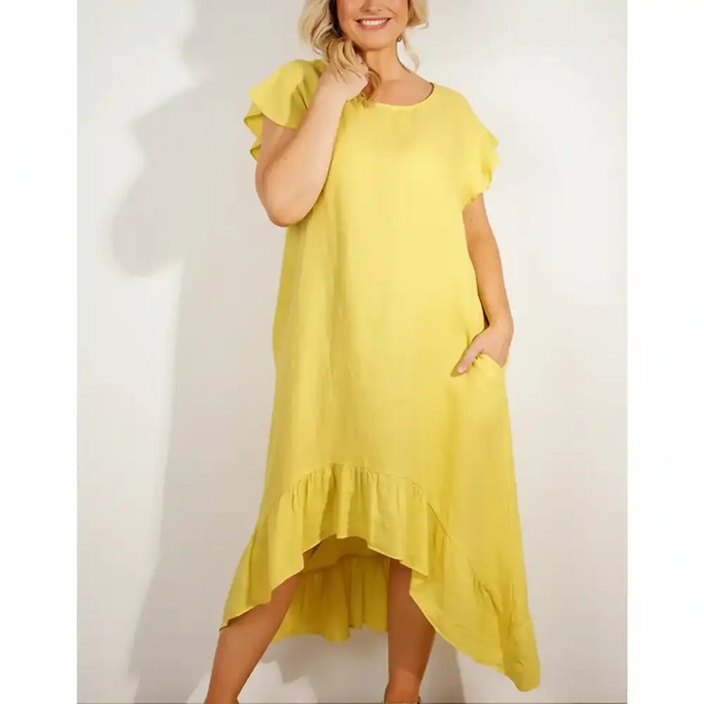 A woman wearing a loose, short-sleeve yellow dress with ruffled shoulders and a ruffled high-low hem, standing against a plain light wall.