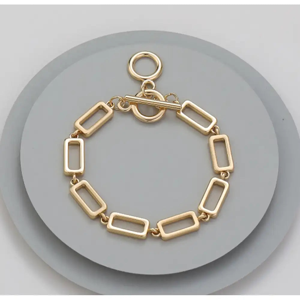 A gold-toned rectangular link bracelet with a toggle clasp, displayed on a round gray stand against a light gray background.
