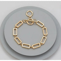 A gold-toned rectangular link bracelet with a toggle clasp, displayed on a round gray stand against a light gray background.