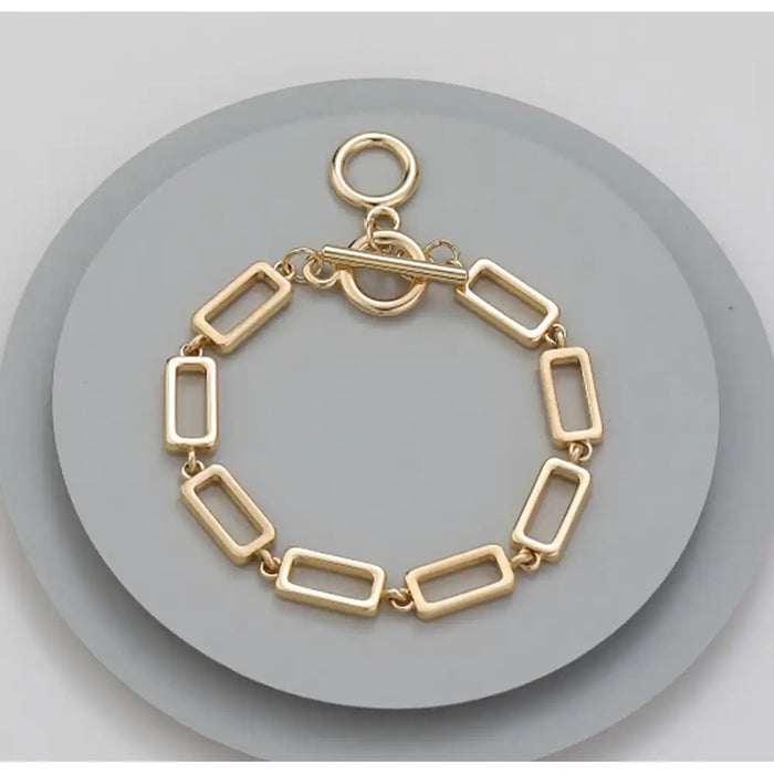 A gold-toned rectangular link bracelet with a toggle clasp, displayed on a round gray stand against a light gray background.