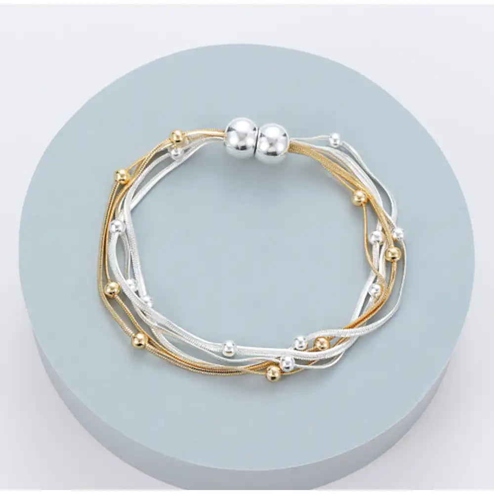 A multi-strand bracelet with gold and silver-tone cords, small metallic beads, and a magnetic silver clasp, displayed on a round light blue pedestal.