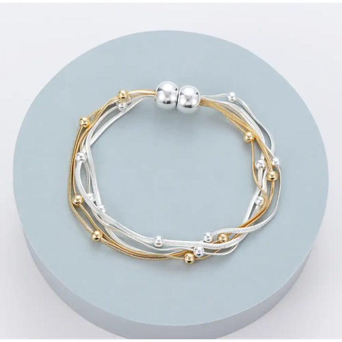 A multi-strand bracelet with gold and silver-tone cords, small metallic beads, and a magnetic silver clasp, displayed on a round light blue pedestal.