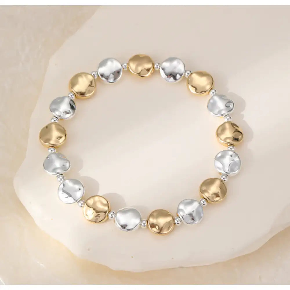 A beaded stretch bracelet featuring alternating hammered gold and silver disc beads, displayed on a light beige stone surface.