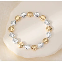A beaded stretch bracelet featuring alternating hammered gold and silver disc beads, displayed on a light beige stone surface.