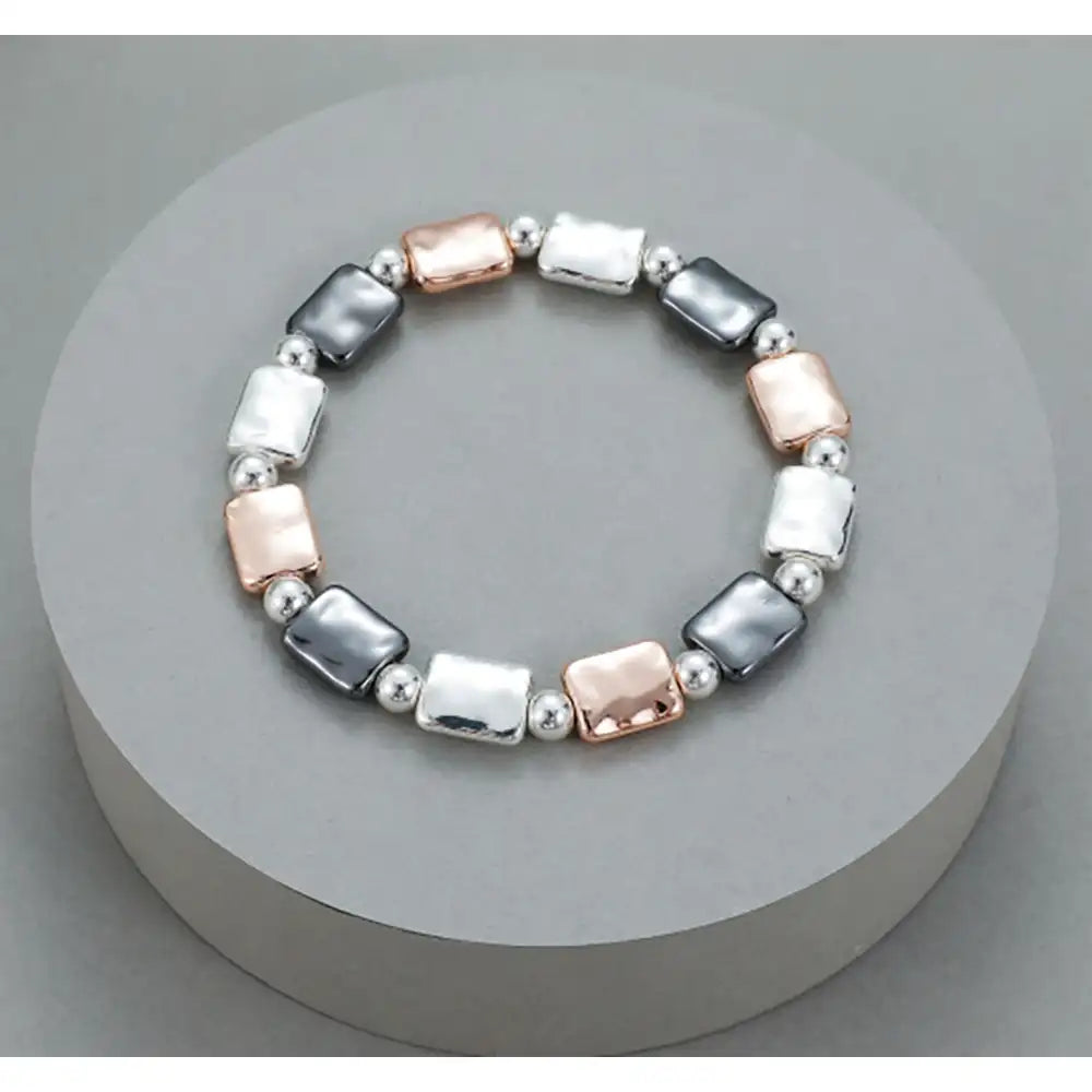 A stretch bracelet featuring alternating polished silver rectangular beads, silver round beads, and rose gold rectangular beads, displayed on a round gray pedestal.