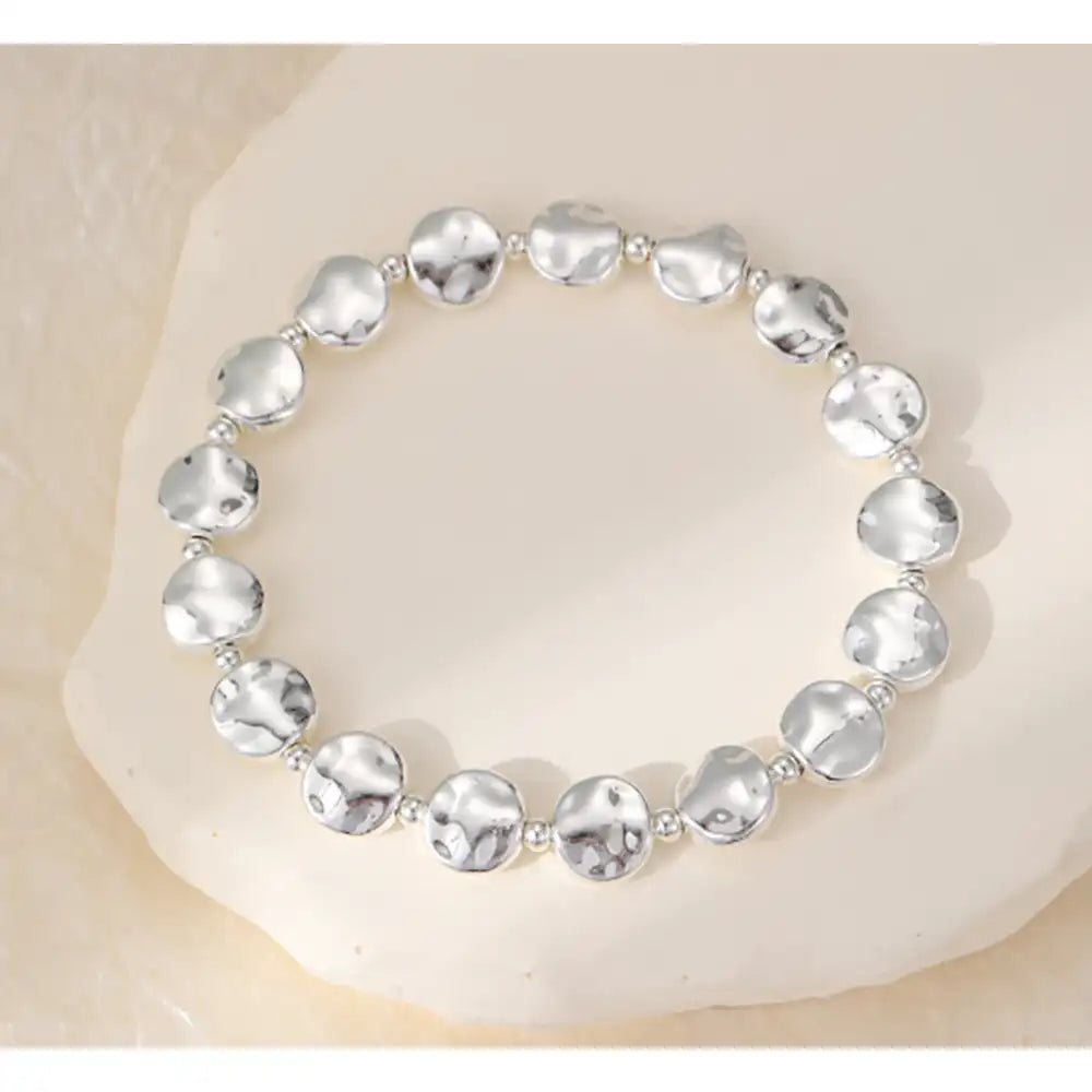 A silver beaded stretch bracelet with irregular, hammered round beads, displayed on a light beige stone surface.
