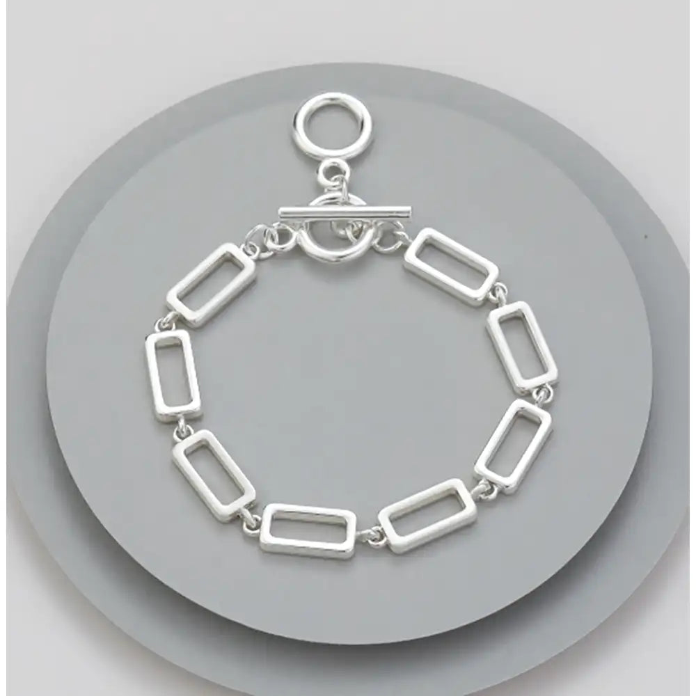 A silver-toned rectangular link bracelet with a toggle clasp, displayed on a round gray stand against a light gray background.